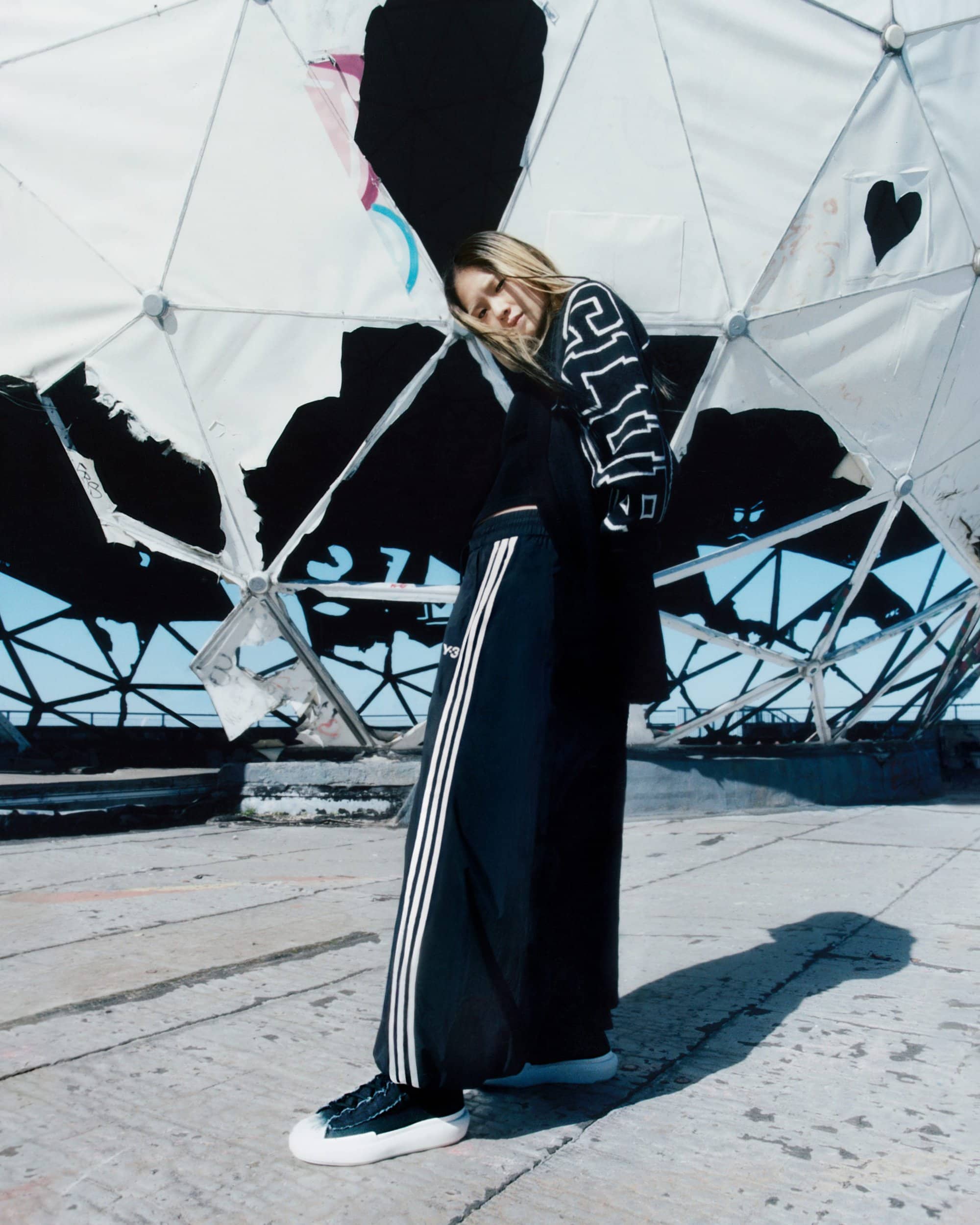 A young woman in a black outfit poses against a weathered dome, her blonde hair contrasting with her sporty-chic look.
