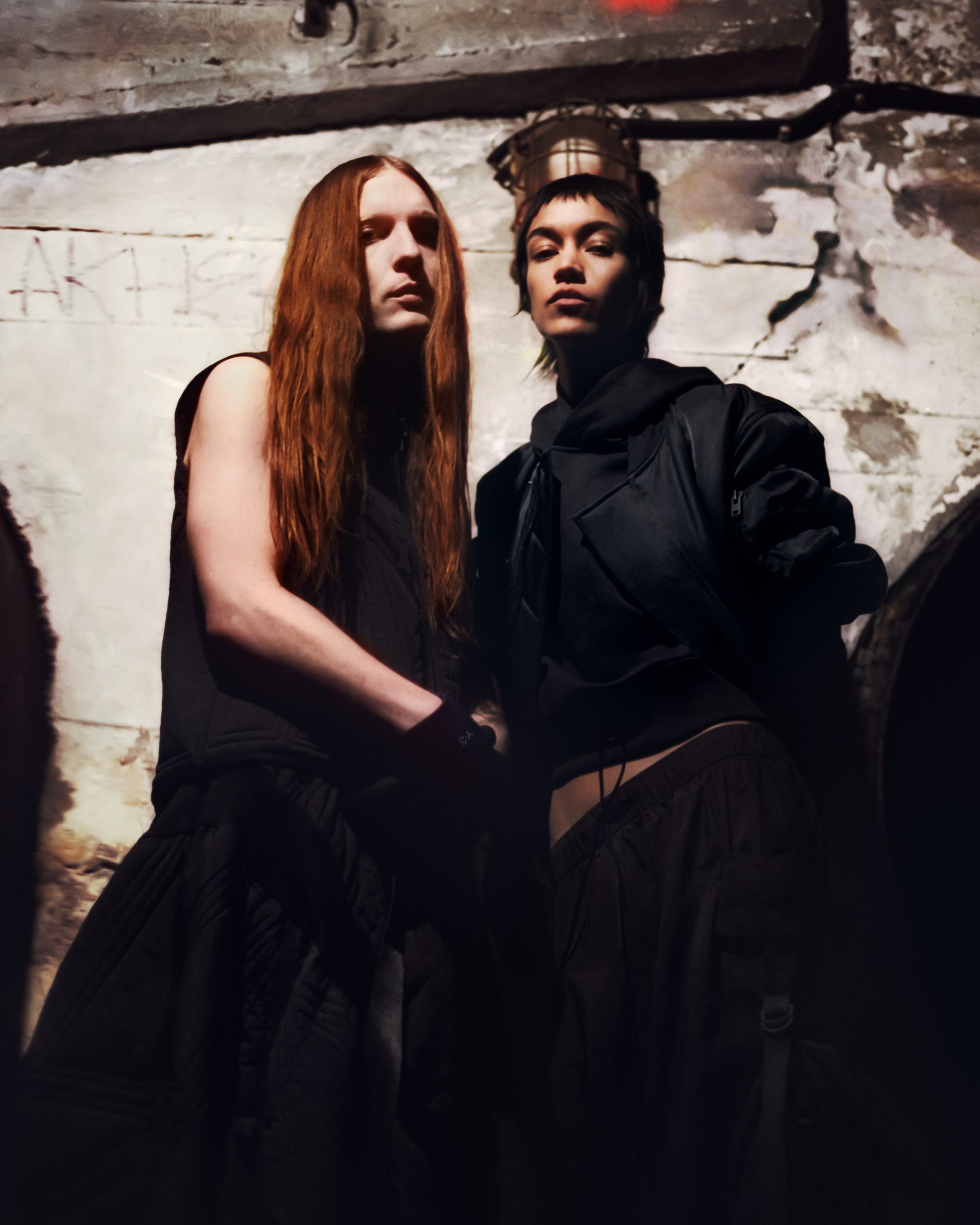 Two models in black clothing pose indoors against a textured wall with graffiti, under dim light, creating an edgy fashion editorial.