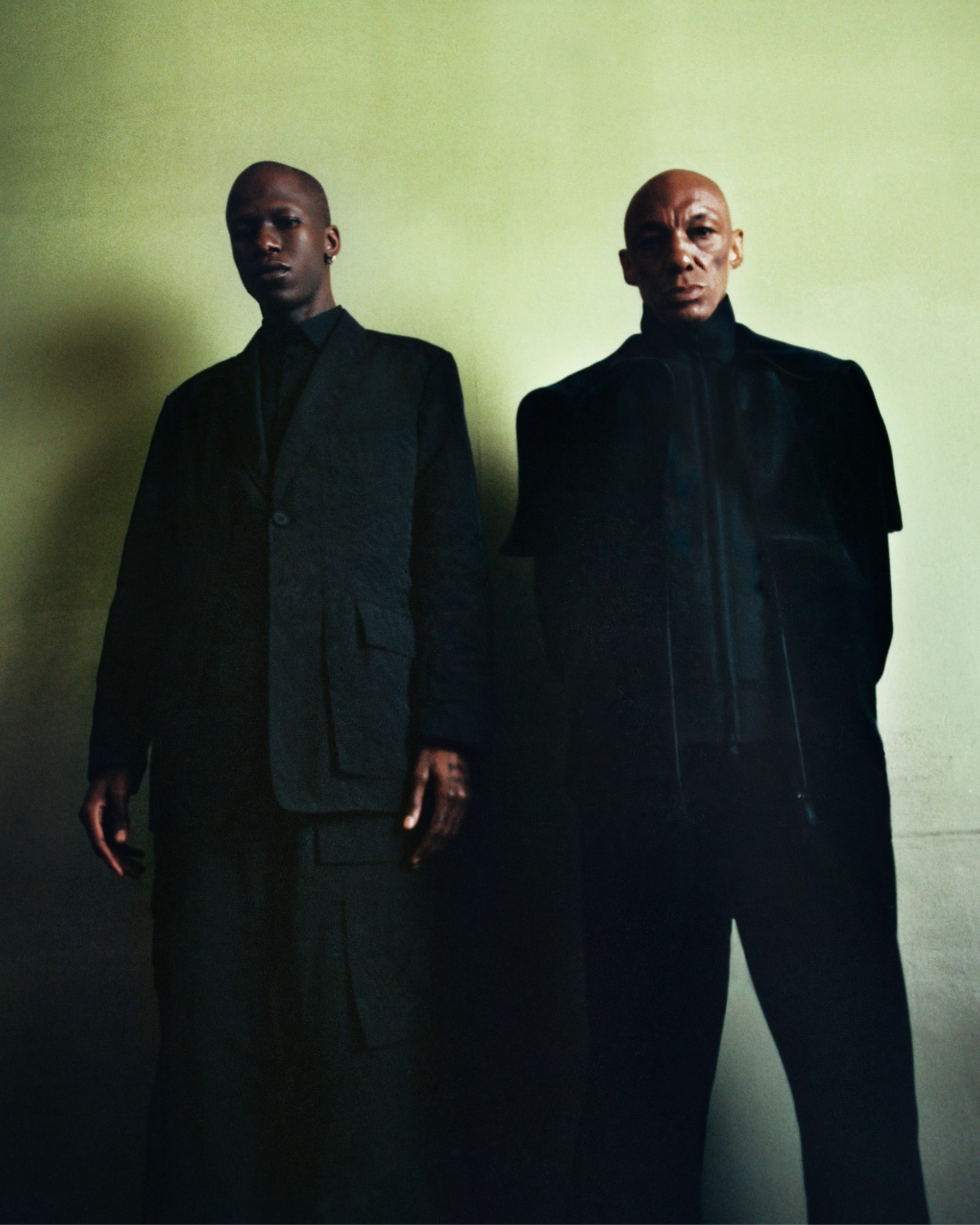 Two bald African-American men in dark suits pose indoors against a neutral wall, looking directly at the camera with serious expressions.