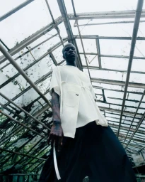 Low-angle shot of a model wearing an off-white t-shirt and black trousers inside a glass greenhouse.