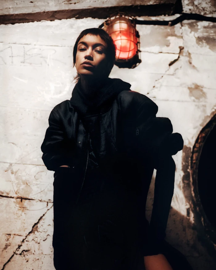 Model in black outerwear standing against a weathered concrete wall beneath a caged wall lamp for Y-3 Fall 2023