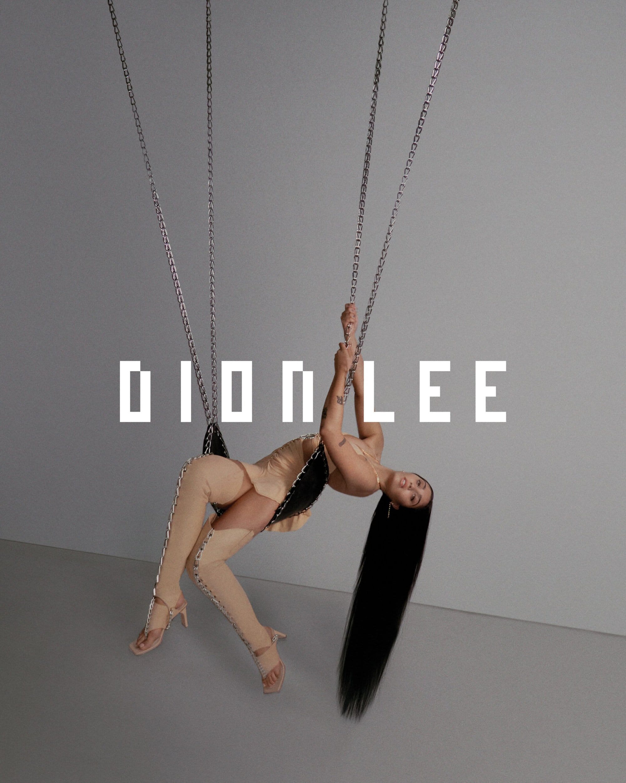 Woman with long hair poses on chains wearing a nude-colored outfit and heels in a studio with soft lighting and neutral background.