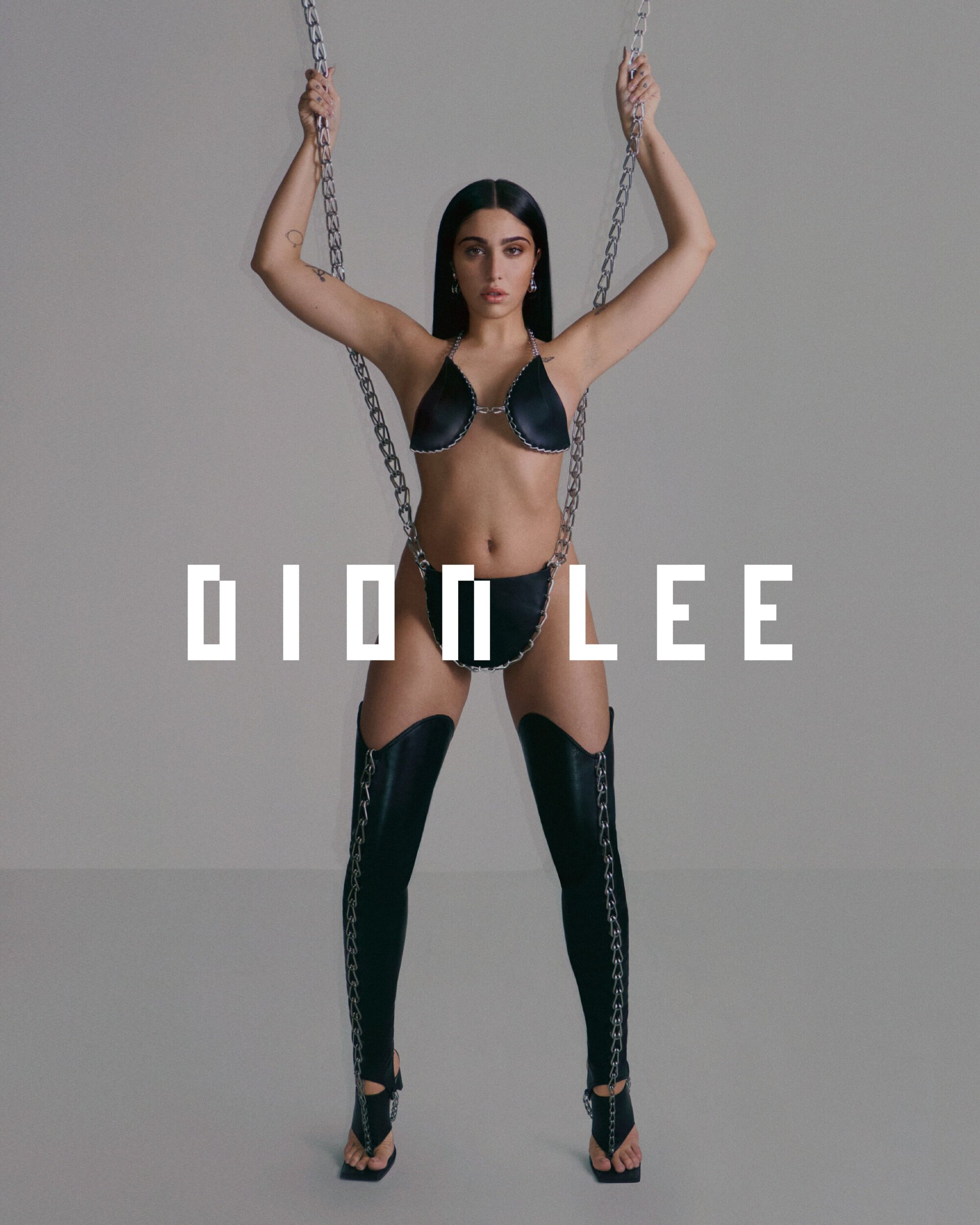 A woman in a black bikini and black thigh-high leggings poses with metal chains against a neutral background, fashion editorial.