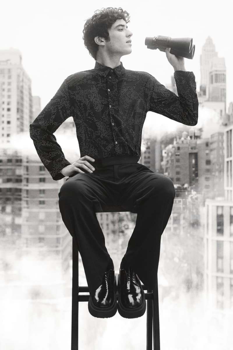 Monochrome portrait of a young adult male seated on a stool, holding binoculars against a blurred cityscape background.