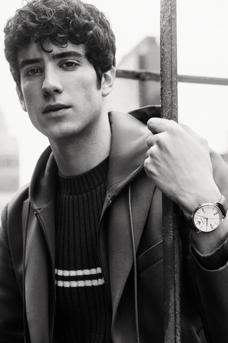 Monochrome portrait of a young adult man with curly hair, wearing a striped sweater and jacket, posing near a metal fence.