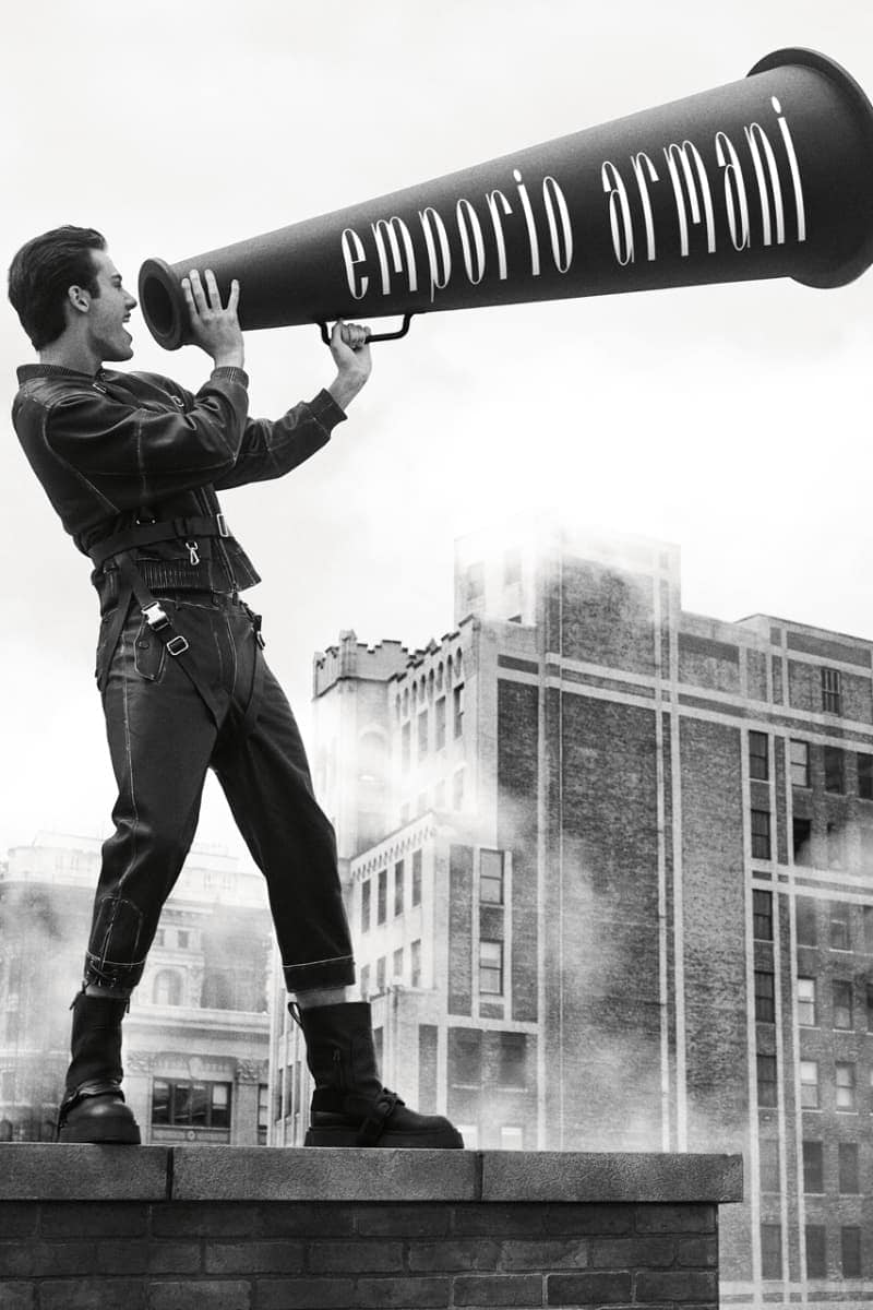 Monochrome image of a man on a rooftop holding an oversized Emporio Armani megaphone against a city and cloudy sky backdrop.