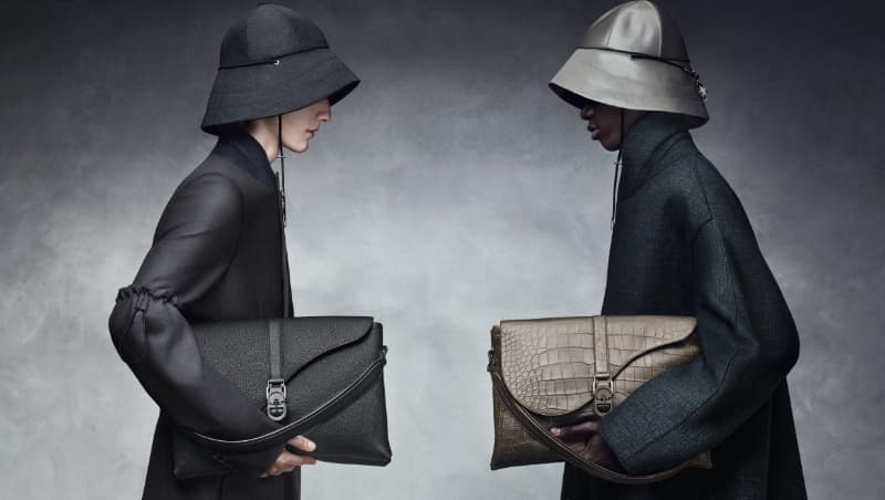 Two fashion models in hats holding handbags in a studio setting with a grey background, showcasing modern style and design.