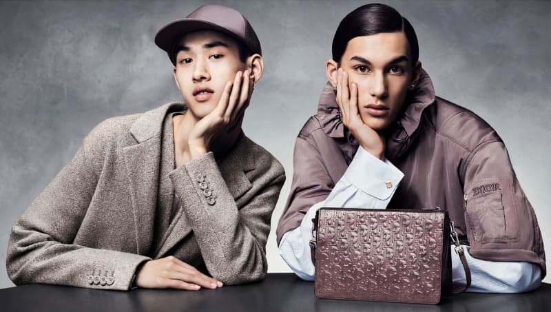 Two people pose with a Dior purse in a studio. One person wears a blazer, and the other is wearing a bomber jacket.