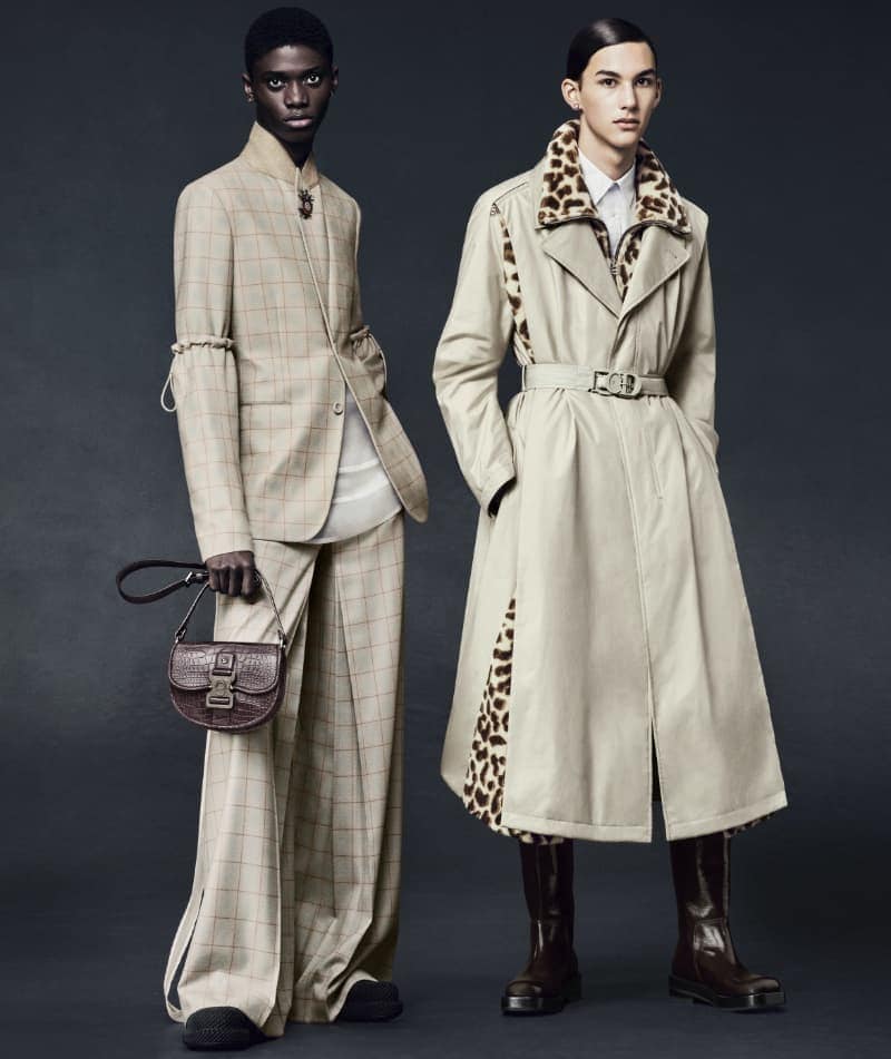 Two fashion models in a studio setting. One wears a beige plaid suit and carries a brown purse. The other wears a trench coat.