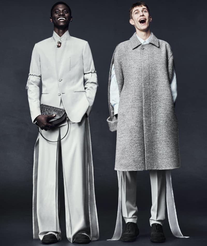 Two male models posing in tailored Dior menswear, one in a light suit holding a saddle bag and one in a grey tweed sleeveless coat.
