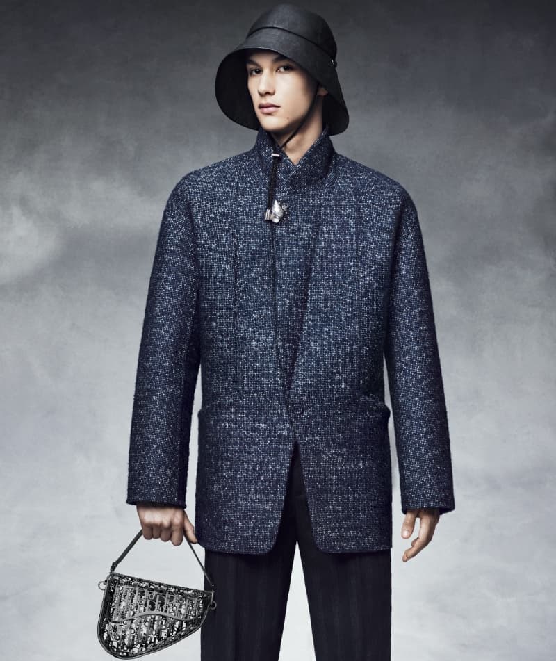 Male model in a grey textured coat and black hat holds a saddle bag against a grey studio backdrop for a fashion editorial.