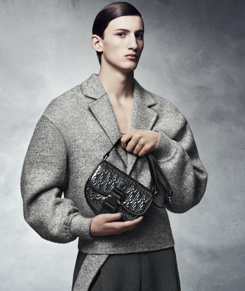 Androgynous person in a grey suit jacket holding a black leather Dior saddle bag with silver hardware against a grey background.