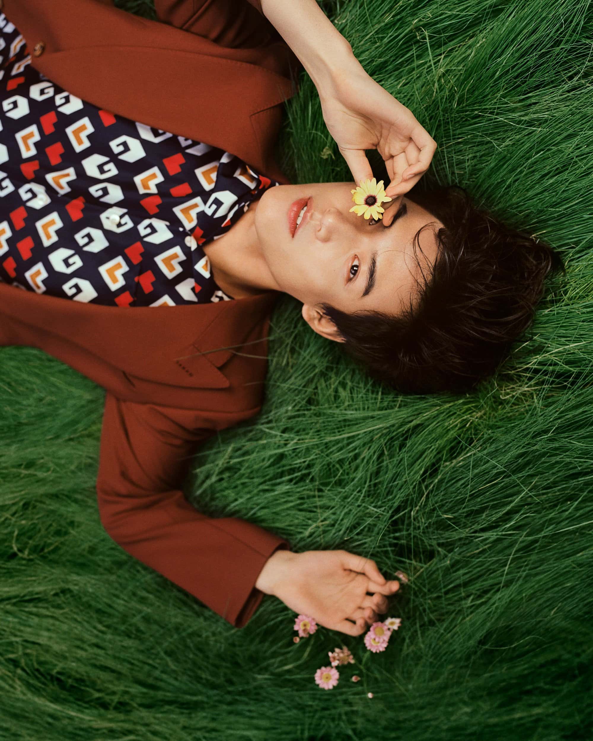 An adult male lies in green grass, holding a flower near his eye, wearing a brown blazer and patterned shirt in an eye-level outdoor shot.