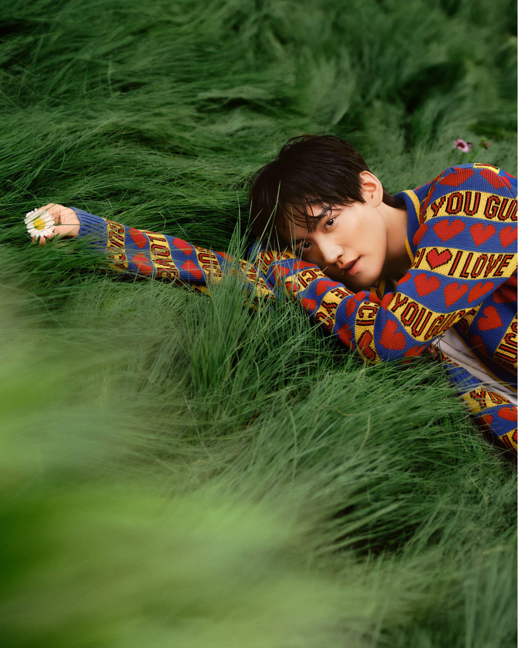 A young adult male lies in a field of tall green grass wearing a patterned knit sweater, holding a daisy in his hand, looking at the camera.