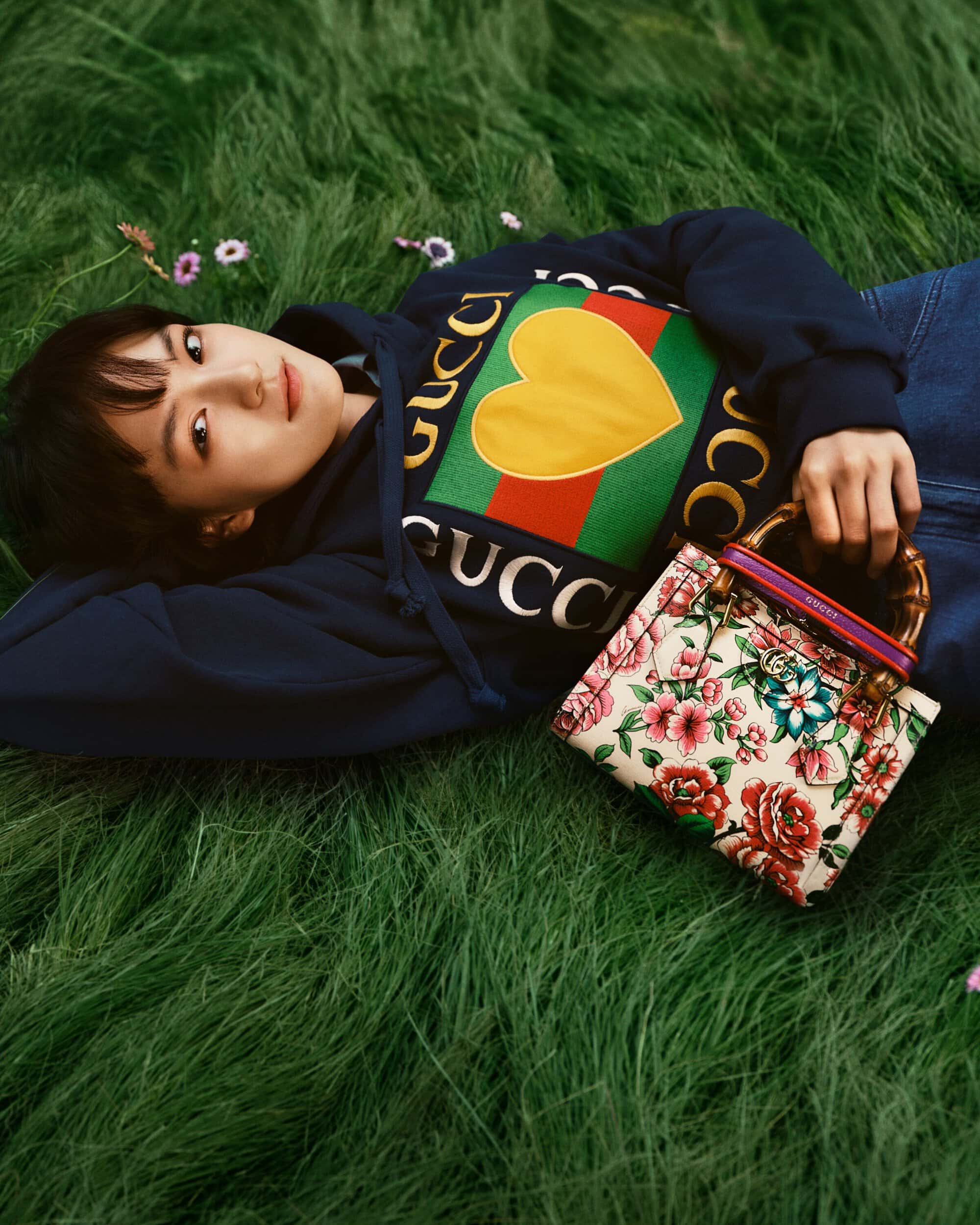 Woman in a hoodie lying in green grass with a Gucci handbag. Fashion shot featuring a person outdoors surrounded by lush greenery.