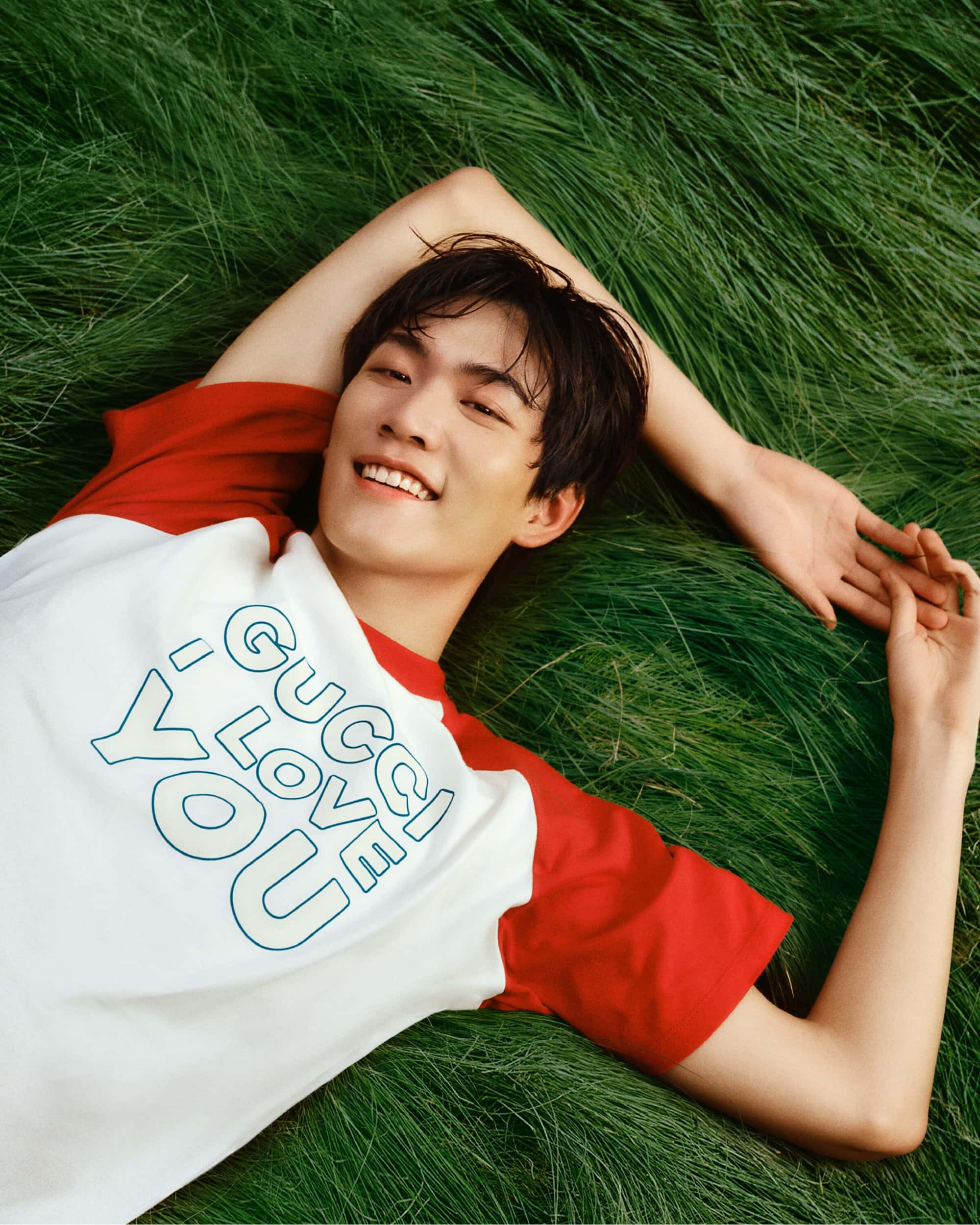 An Asian male model lies on bright green grass in a white and red Gucci t-shirt, smiling with his arms outstretched in a beauty shot.