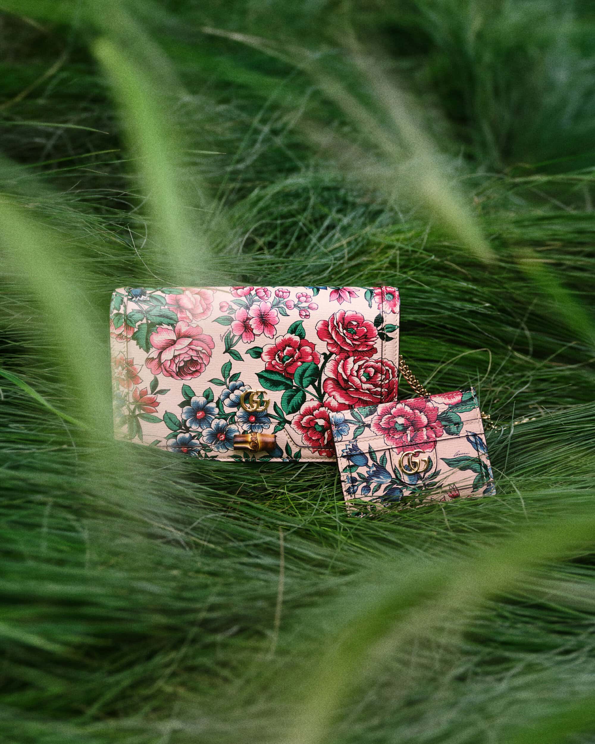 Floral-patterned Gucci bags set in green grass. Close up of designer purses. Luxury accessories with pink, red, and blue flower designs.