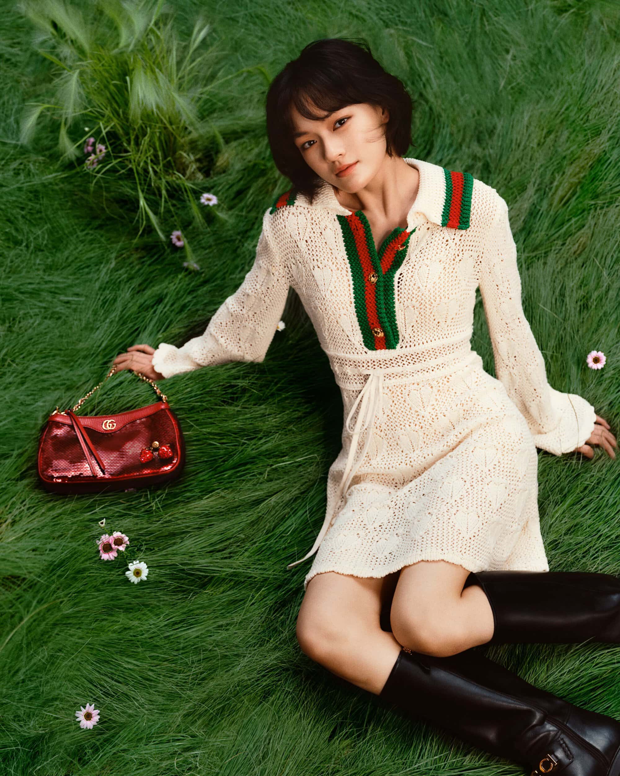 Woman in a cream-colored crocheted dress and black boots lies on lush green grass with a red shoulder bag, surrounded by small white flowers.