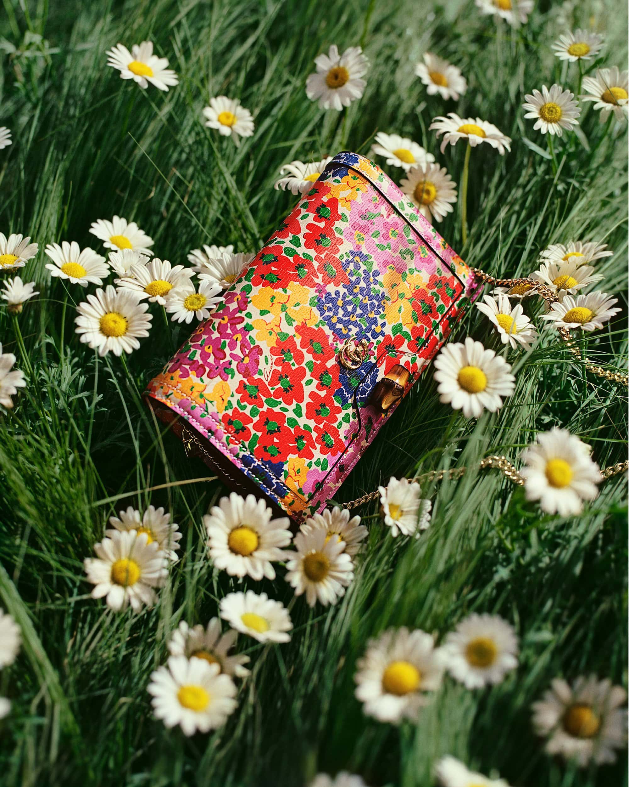 Close-up of a floral-print purse with a gold chain strap laying in green grass with scattered daisy flowers, shot in natural light.