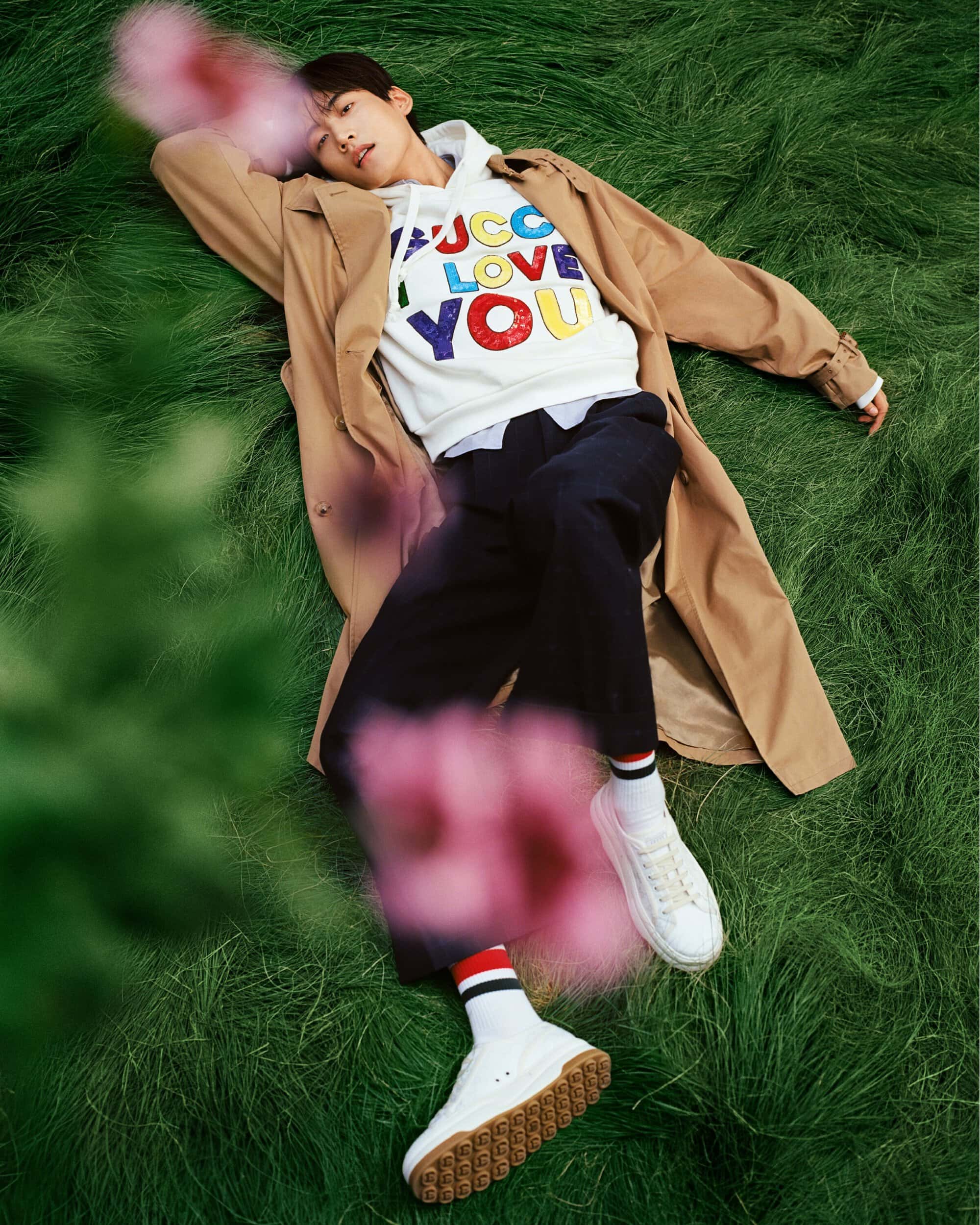 An Asian man lies on his side in green grass, wearing a tan coat, white hoodie with colorful letters, black pants, and white sneakers.