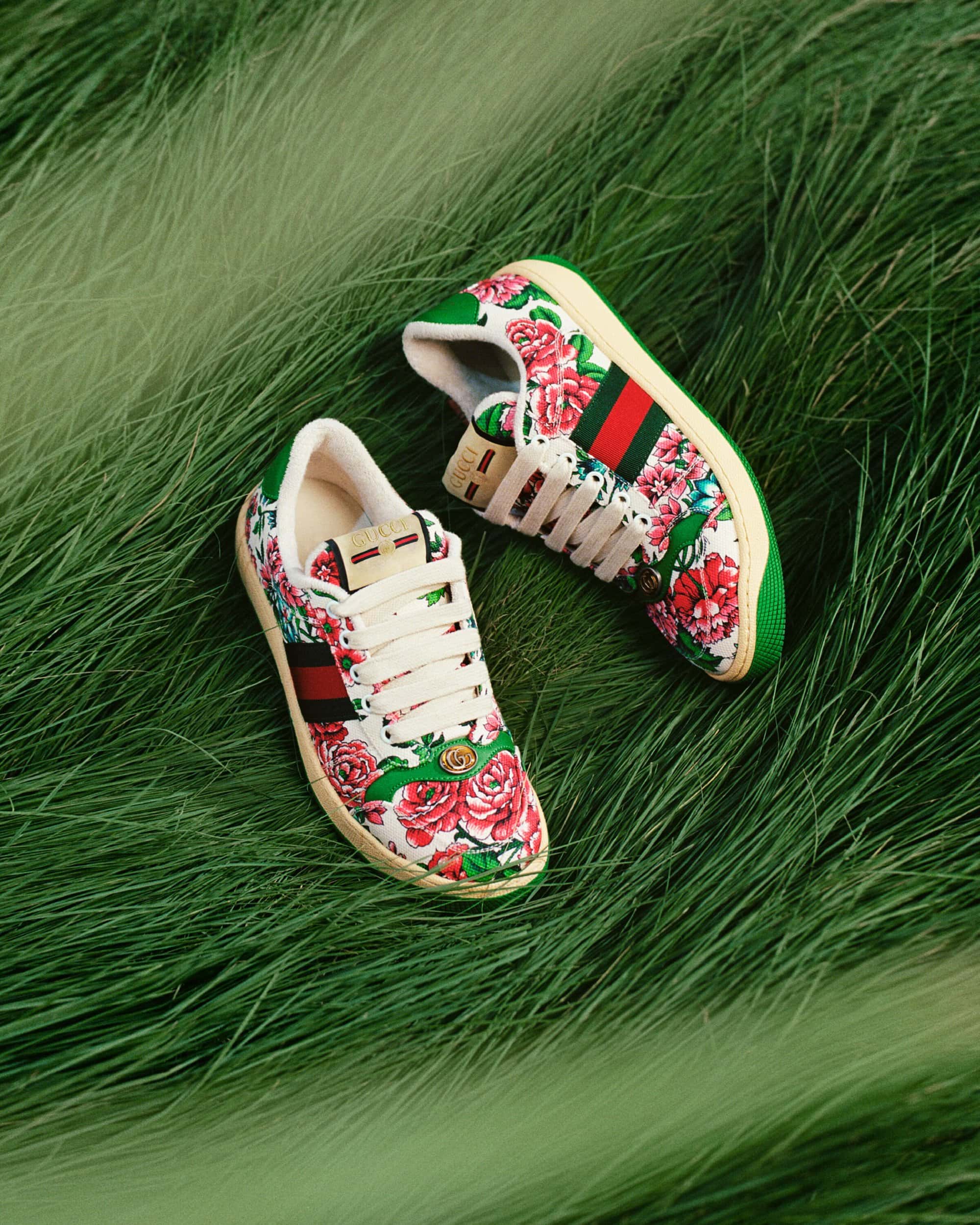 Two floral Gucci sneakers with red and green stripes and white laces resting on a bed of green grass.