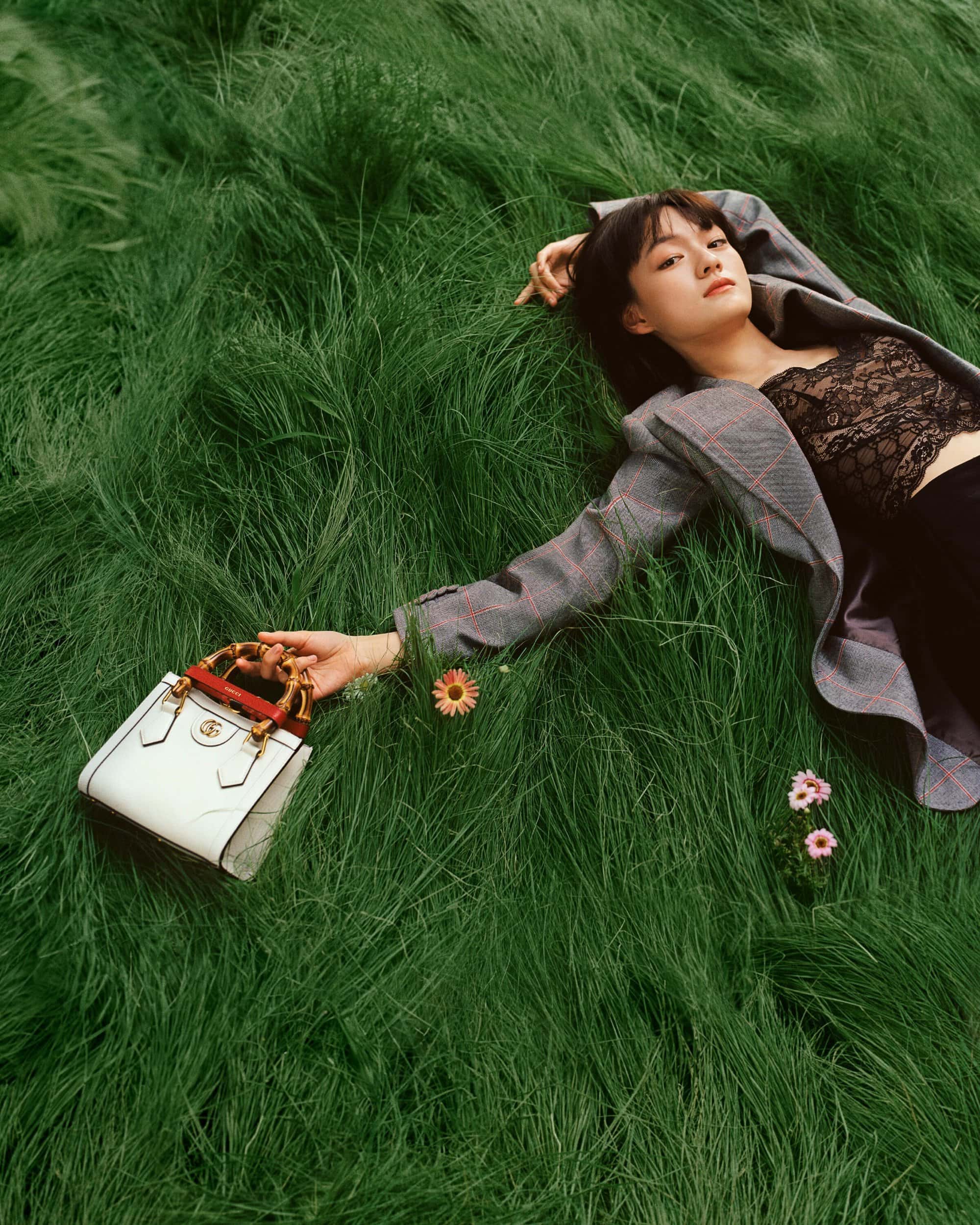 A woman lies on green grass wearing a blazer, with a purse, surrounded by greenery and small flowers in an outdoor setting.