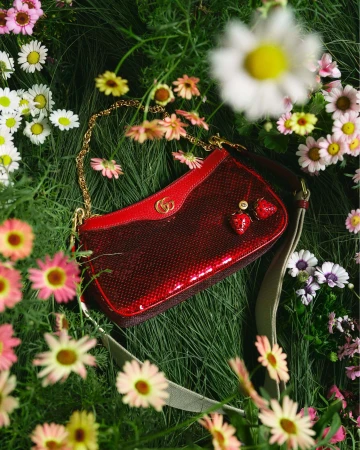 A red Gucci handbag with strawberry charms rests on green grass surrounded by colorful daisies in an outdoor setting, showcasing a floral design.