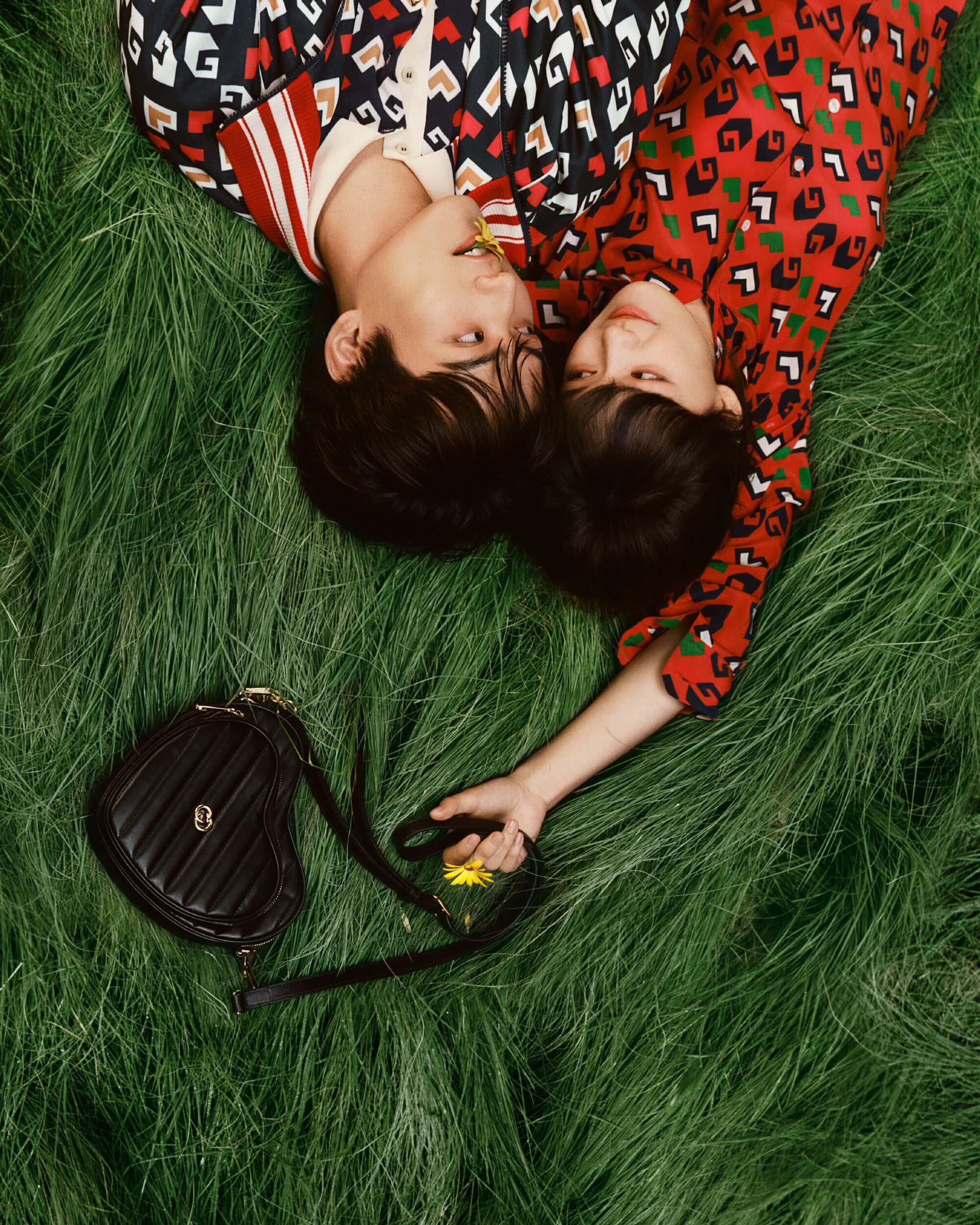 Overhead shot of a stylish couple lying on green grass with a black heart-shaped purse, holding yellow flowers in an outdoor setting.