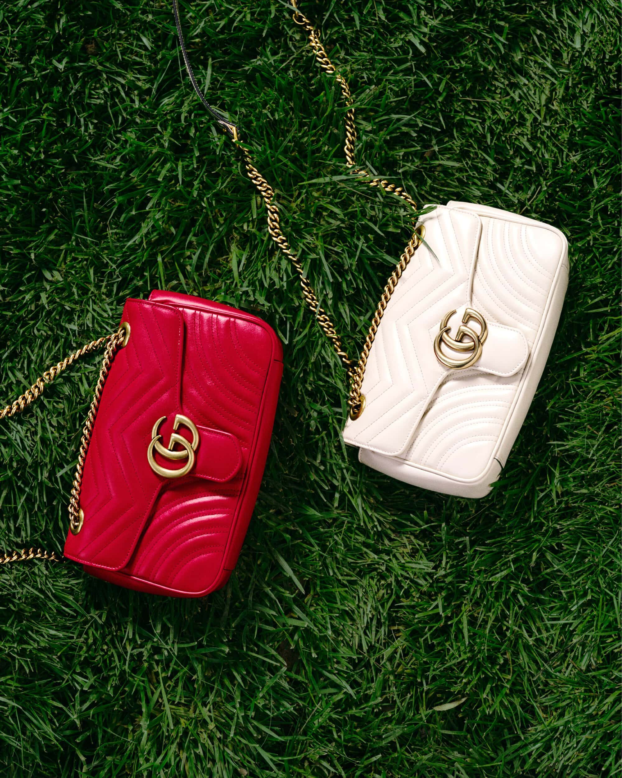 Two Gucci Marmont handbags, one red and one white, sit on a bed of vibrant green grass with their gold chain straps displayed.