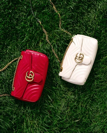 Two Gucci Marmont handbags, one red and one white, sit on a bed of vibrant green grass with their gold chain straps displayed.
