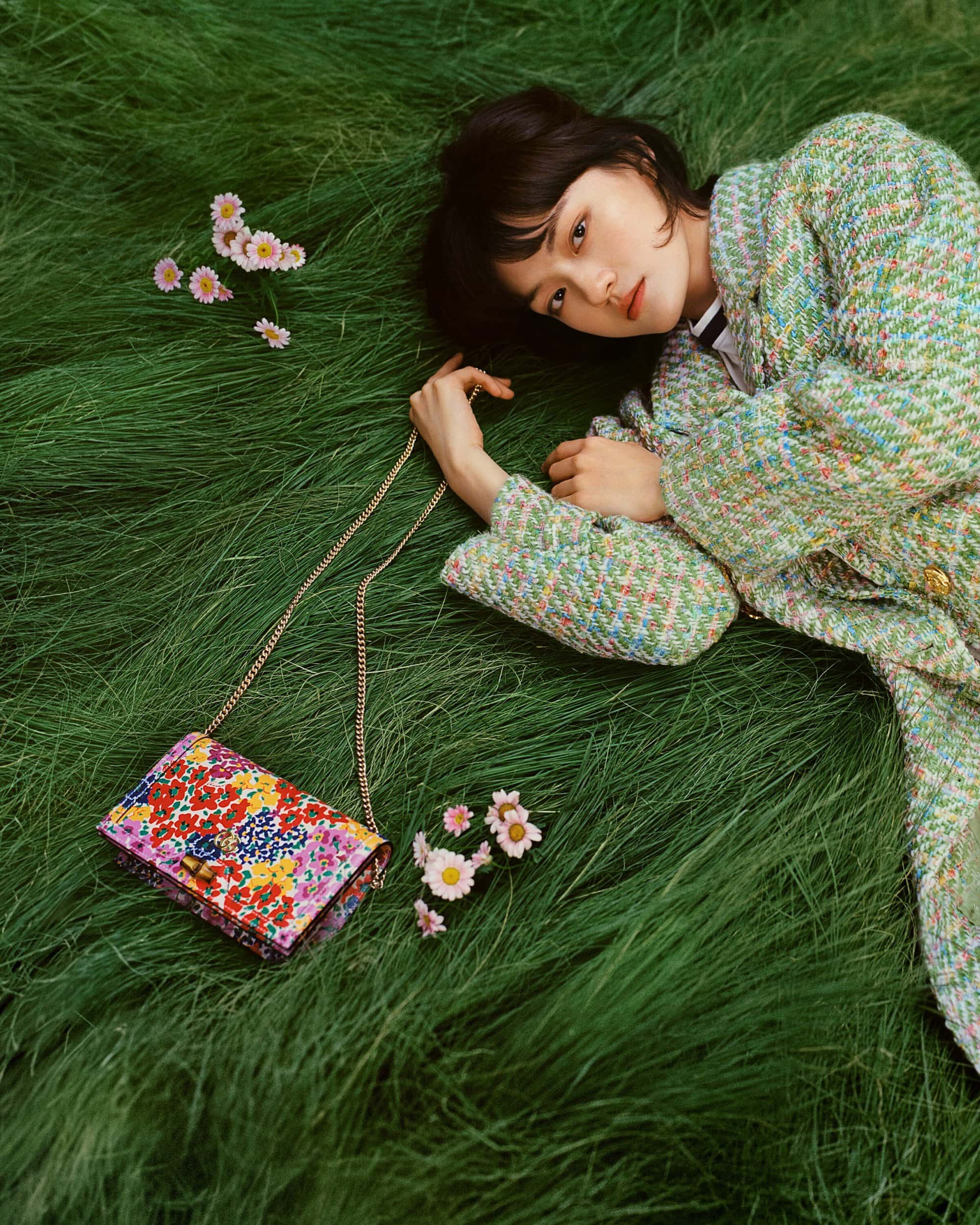 Young woman with short hair, wearing a tweed jacket, laying in green grass with a colorful floral purse and daisies scattered nearby.