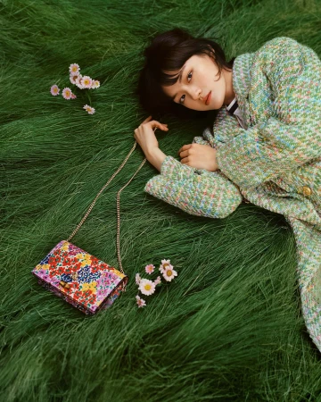 Young woman with short hair, wearing a tweed jacket, laying in green grass with a colorful floral purse and daisies scattered nearby.
