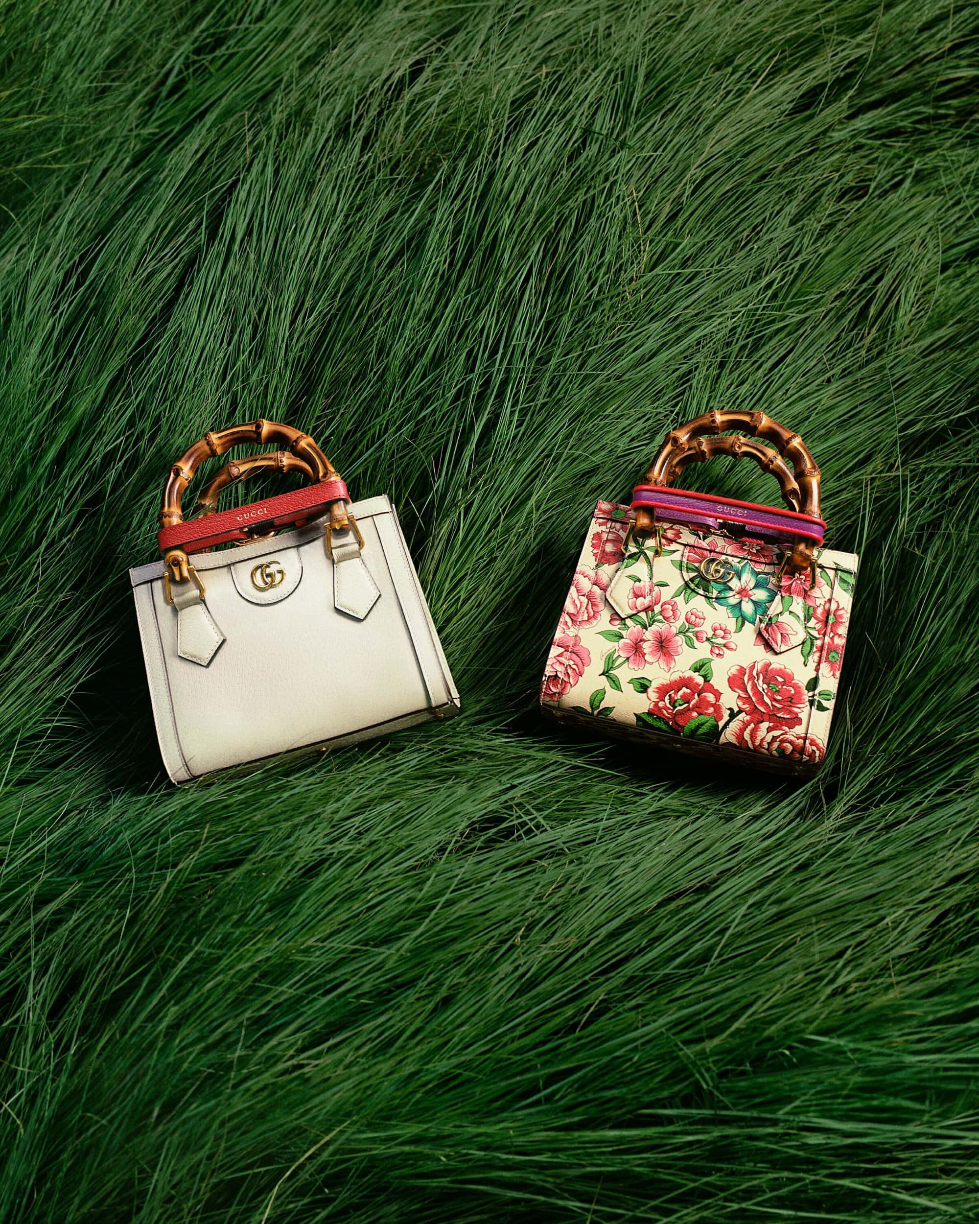 Two Gucci bags sit on a bed of green grass. One bag is white, and the other is floral patterned with bamboo handles.