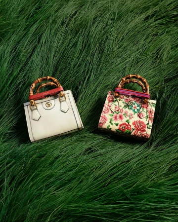Two Gucci bags sit on a bed of green grass. One bag is white, and the other is floral patterned with bamboo handles.