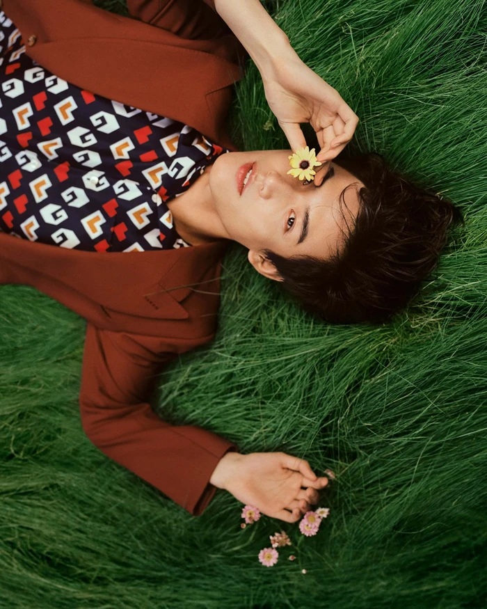A person lying in lush green grass holding a small yellow flower over their eye, wearing a brown suit and patterned shirt.