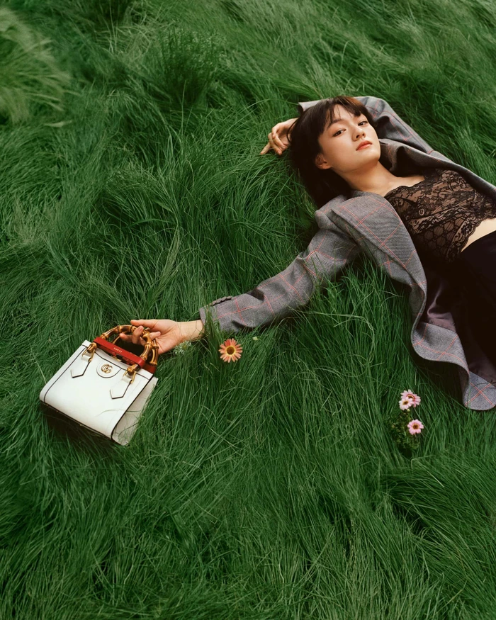Wen Qi reclining in tall green grass holding a white Gucci handbag with a bamboo handle while wearing a grey plaid blazer.