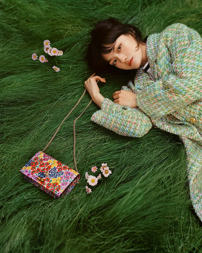 Woman in tweed jacket lying in green grass beside a floral handbag and pink daisies