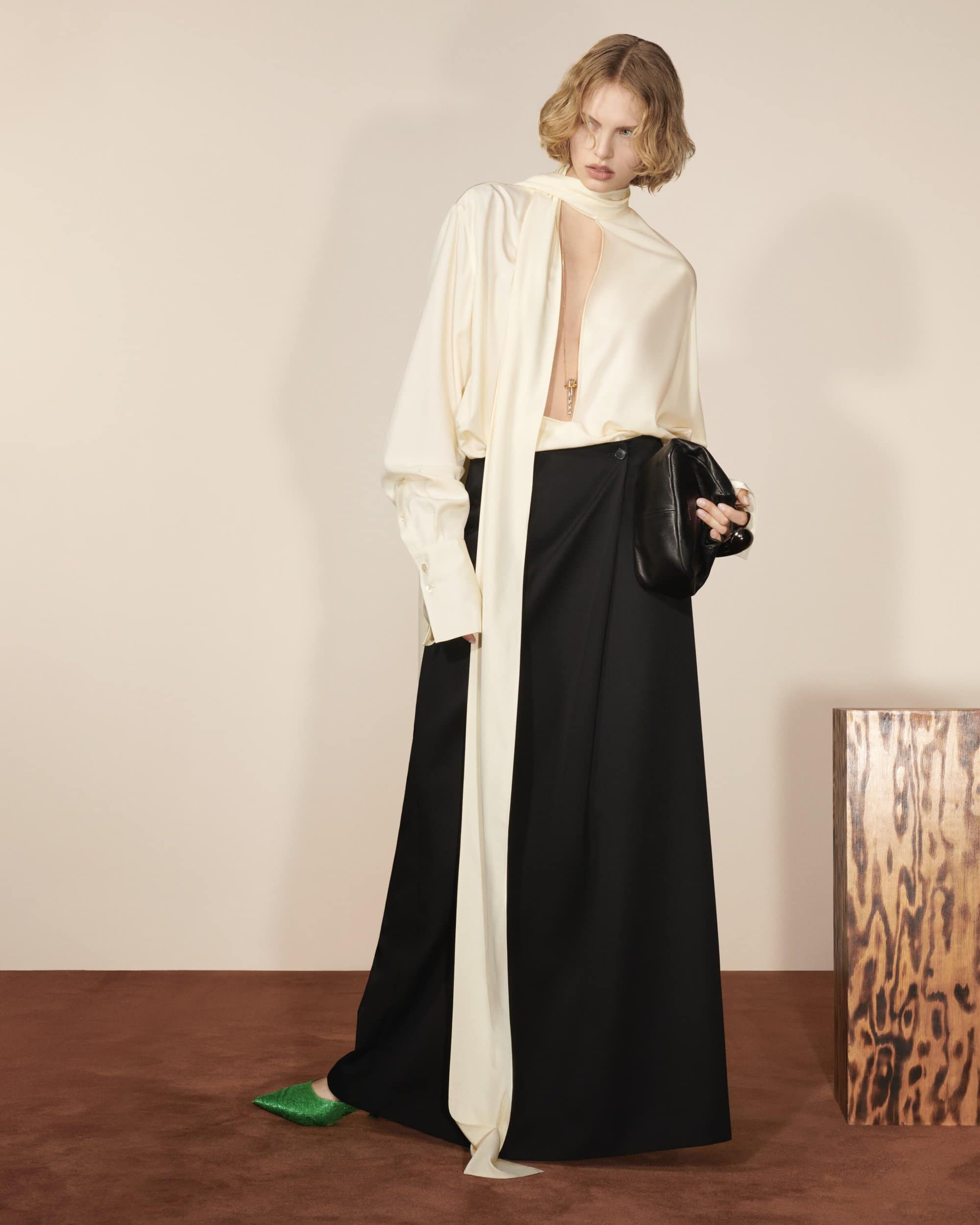 A woman in a cream blouse with a long tie, a black skirt, and green shoes poses indoors against a neutral backdrop with a wooden accent table.