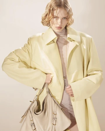 Fashionable woman posing indoors wearing a yellow coat and holding a beige purse, showcasing contemporary style in a studio setting.