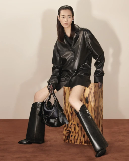 Fashion shot of an Asian female model in an all-black leather outfit, posing with a purse on a wooden block in a studio.