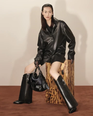 Fashion shot of an Asian female model in an all-black leather outfit, posing with a purse on a wooden block in a studio.
