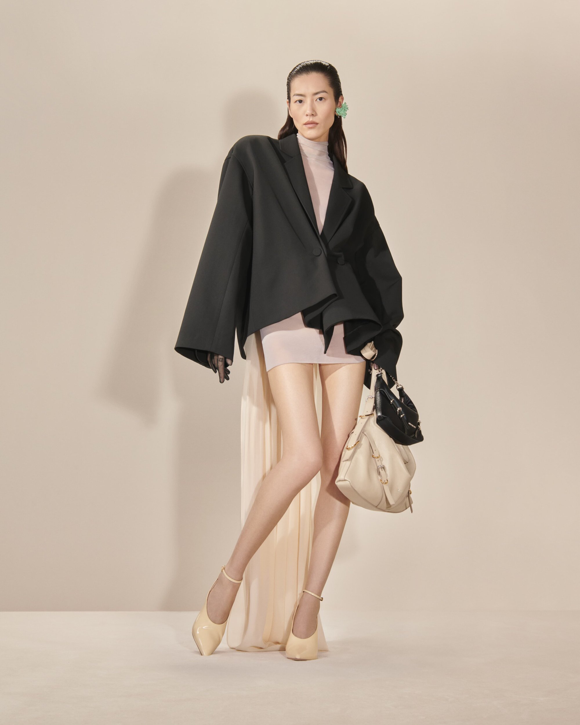 An Asian woman stands confidently in a studio, wearing a black blazer, tan mini dress, beige heels, and carrying a beige handbag.