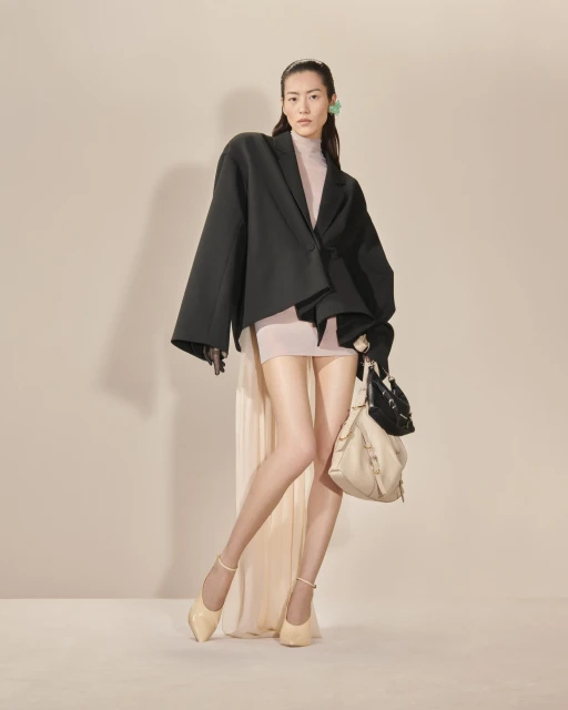 An Asian woman stands confidently in a studio, wearing a black blazer, tan mini dress, beige heels, and carrying a beige handbag.