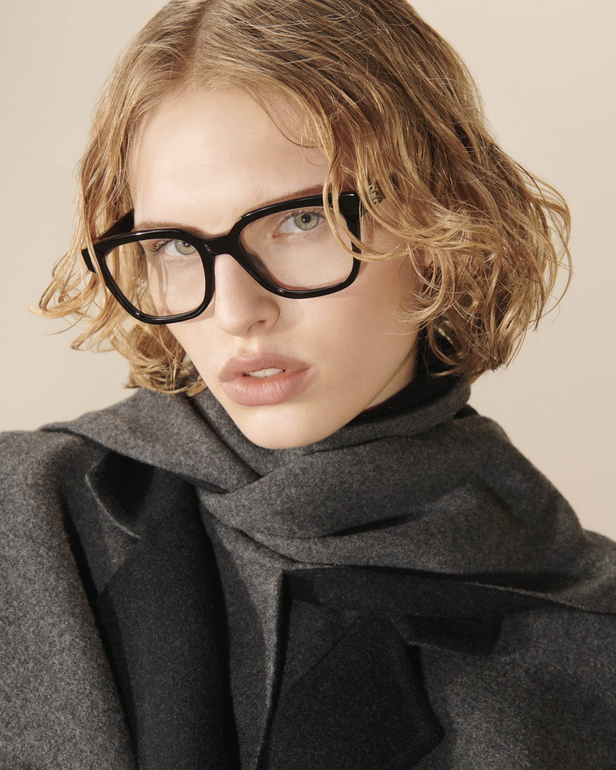 Close-up studio shot of a blonde woman with glasses, wearing a grey scarf. Fashion and portrait photography, featuring modern eyewear.