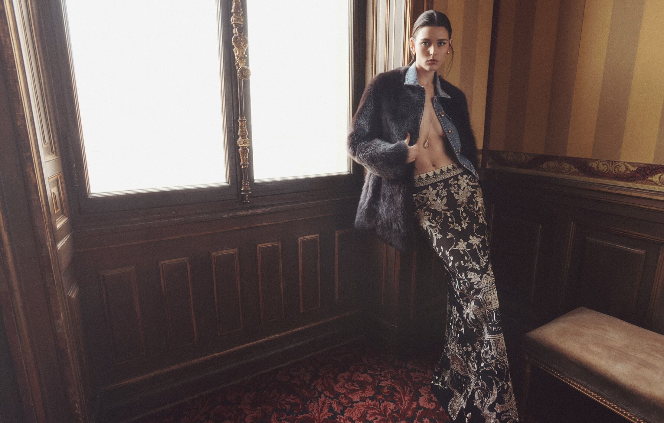 Indoor fashion shot featuring a slender adult woman in a fur coat, denim shirt, and floral pants leaning against a dark wood paneled wall.