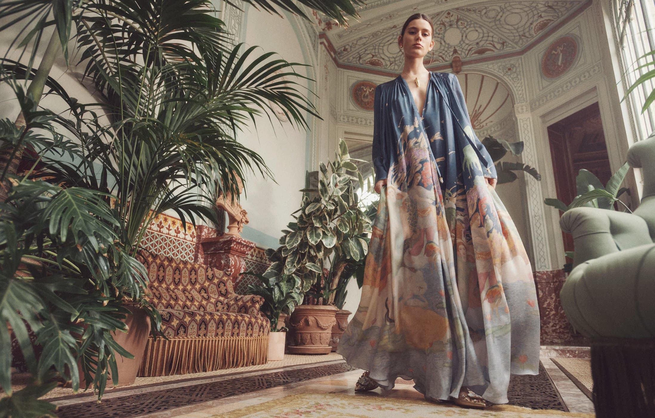 A woman in a patterned dress poses in a bright indoor setting with plants and decorative furniture, showcasing fashion and interior design.