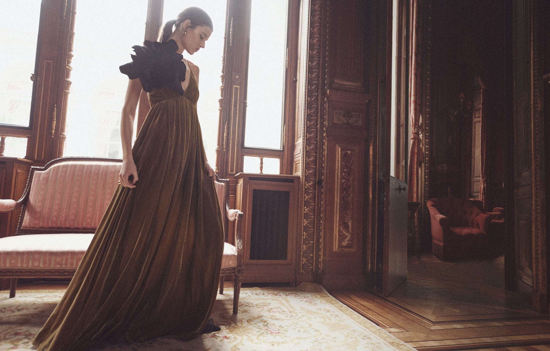 A woman in a long olive green dress with a black feathered epaulette poses elegantly in an ornate room with large windows and vintage furniture.