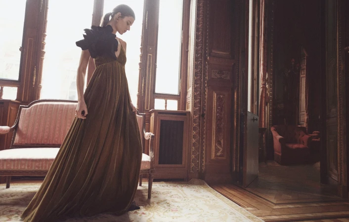 A woman in a long olive green dress with a black feathered epaulette poses elegantly in an ornate room with large windows and vintage furniture.