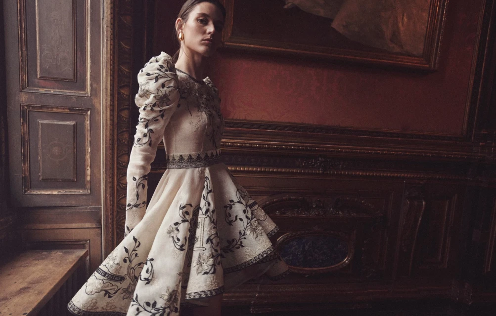 Fashion editorial shot of a female model wearing a floral dress in an indoor setting with wood paneling and artwork in the background.