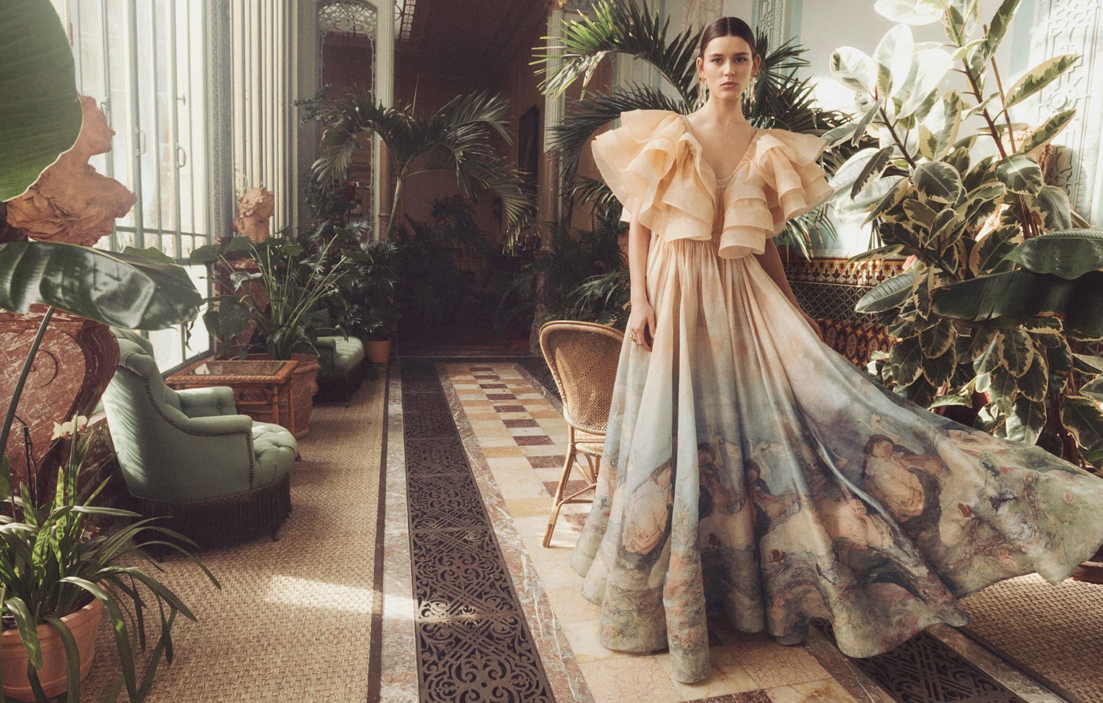 Woman in ruffled beige gown with mural train in bright sunroom filled with lush potted plants and vintage furniture and tiled floor.